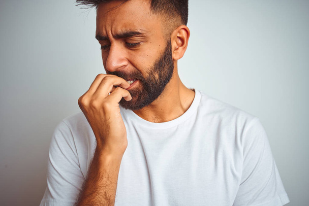 Jonge Indiase man dragen t-shirt staande over geïsoleerde witte achtergrond op zoek gestrest en nerveus met handen op de mond bijten nagels. Angst probleem. - Foto, afbeelding