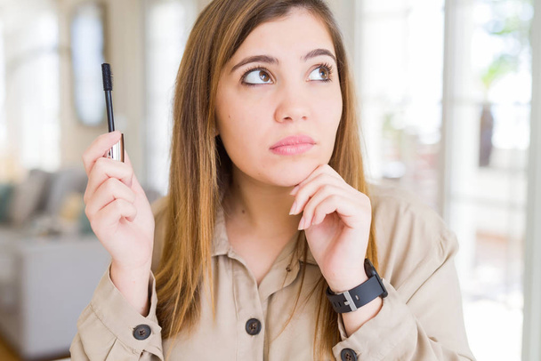 Beautiful young woman putting lash mascara on eyelashes serious face thinking about question, very confused idea - 写真・画像