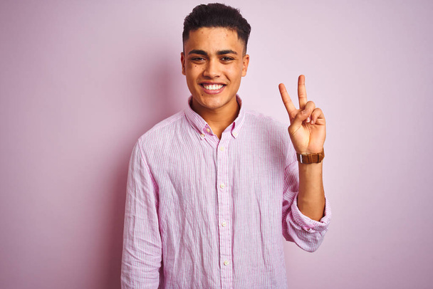 孤立したピンクの背景の上に立つシャツを着た若いブラジル人男性は、自信を持って幸せに微笑みながら、指ナンバー2を示し、指を上げている. - 写真・画像