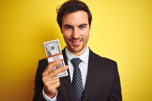 Young handsome businessman wearing suit holding dollars over isolated yellow background with a happy face standing and smiling with a confident smile showing teeth - Photo, Image