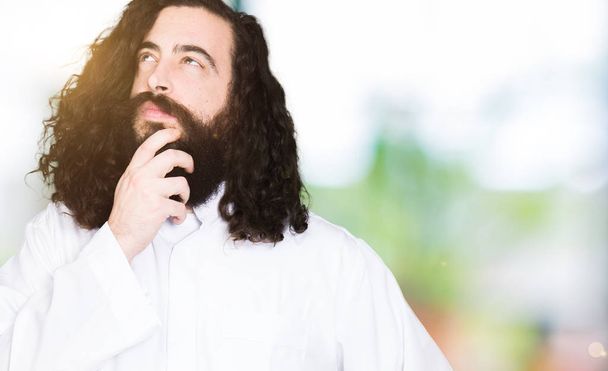 Man wearing Jesus Christ costume with hand on chin thinking about question, pensive expression. Smiling with thoughtful face. Doubt concept. - Photo, Image