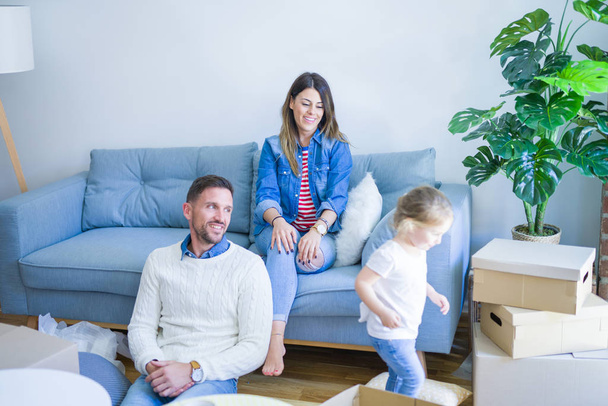 schöne Familie, Eltern sitzen auf dem Sofa und trinken Kaffee und schauen seinem Kind zu, wie es im neuen Zuhause um Pappkartons spielt - Foto, Bild