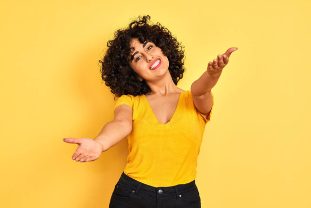 Mujer árabe joven con el pelo rizado usando camiseta de pie sobre el fondo amarillo aislado mirando a la cámara sonriendo con los brazos abiertos para el abrazo. Expresión alegre abrazando la felicidad
. - Foto, imagen