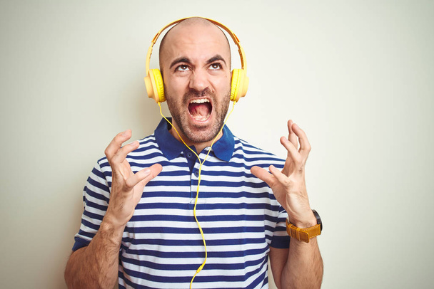 Young man listening to music wearing yellow headphones over isolated background crazy and mad shouting and yelling with aggressive expression and arms raised. Frustration concept. - Fotografie, Obrázek