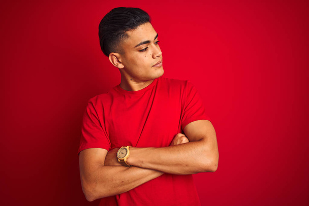 Young brazilian man wearing t-shirt standing over isolated red background looking to the side with arms crossed convinced and confident - Foto, Bild