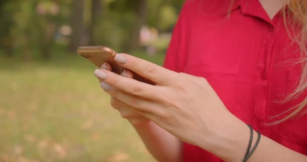 Closeup portrait of young blonde long haired pretty female using phone sitting on grass in park outdoors - Filmagem, Vídeo