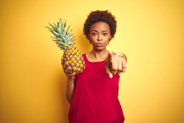 Mujer afroamericana sosteniendo piña tropical sobre fondo aislado amarillo señalando con el dedo a la cámara y a usted, signo de mano, gesto positivo y seguro desde el frente
 - Foto, imagen