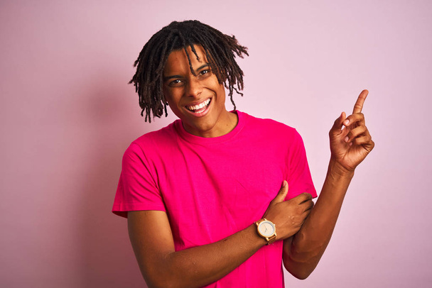 Afro american man with dreadlocks wearing t-shirt standing over isolated pink background with a big smile on face, pointing with hand and finger to the side looking at the camera. - Zdjęcie, obraz