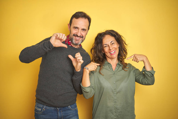 Beautiful middle age couple over isolated yellow background looking confident with smile on face, pointing oneself with fingers proud and happy. - Photo, Image