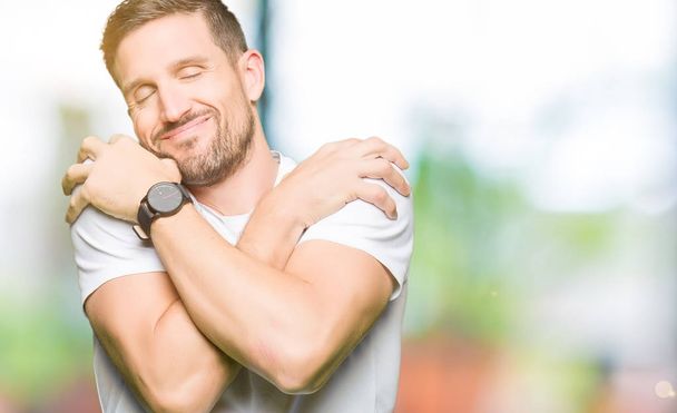 Handsome man wearing casual white t-shirt Hugging oneself happy and positive, smiling confident. Self love and self care - Фото, изображение