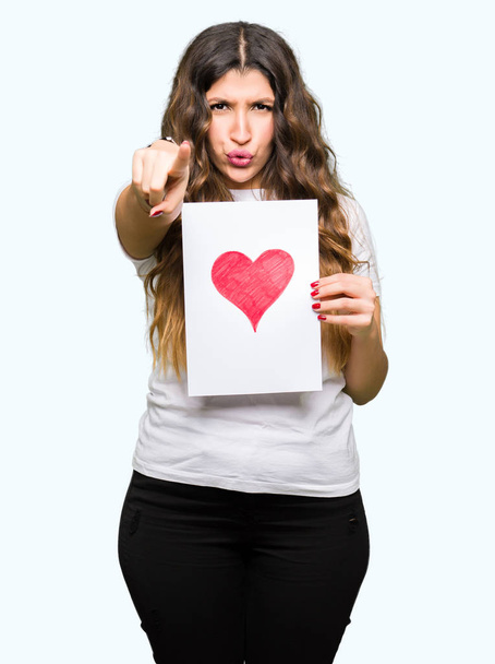Young adult woman holding card with red heart pointing with finger to the camera and to you, hand sign, positive and confident gesture from the front - Фото, изображение