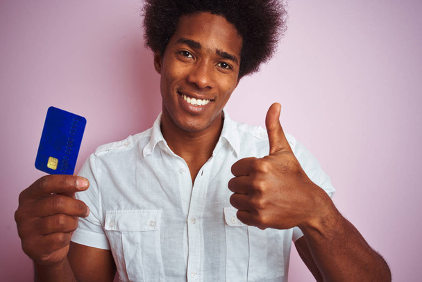 Afro amerikkalainen asiakas mies tilalla luottokortti seisoo yli eristetty vaaleanpunainen tausta tyytyväinen iso hymy tekee ok merkki, peukalo ylös sormilla, erinomainen merkki
 - Valokuva, kuva