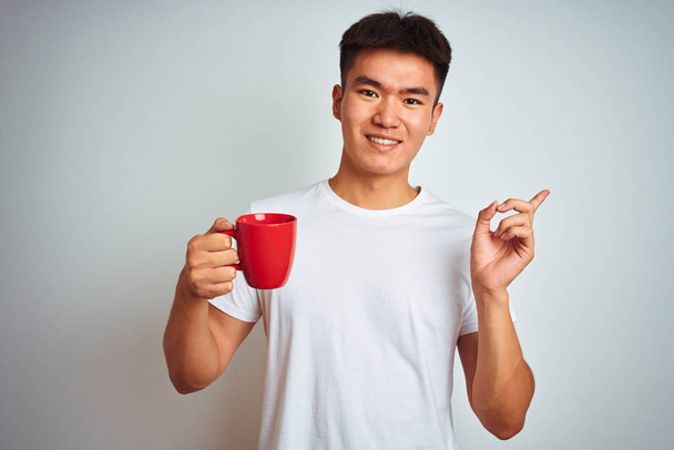 Asian chinese man holding red cup of coffee standing over isolated white background very happy pointing with hand and finger to the side - Фото, изображение