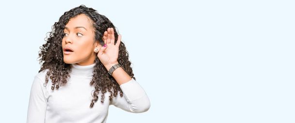 Jonge mooie vrouw met krullend haar dragen coltrui trui glimlachen met hand over oor luisteren een hoorzitting om geruchten of roddels. Doofheid concept. - Foto, afbeelding