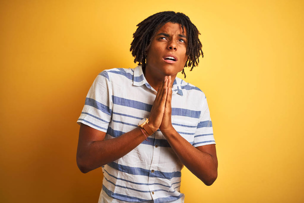 Afro american man with dreadlocks wearing striped shirt over isolated yellow background begging and praying with hands together with hope expression on face very emotional and worried. Asking for forgiveness. Religion concept. - Photo, Image