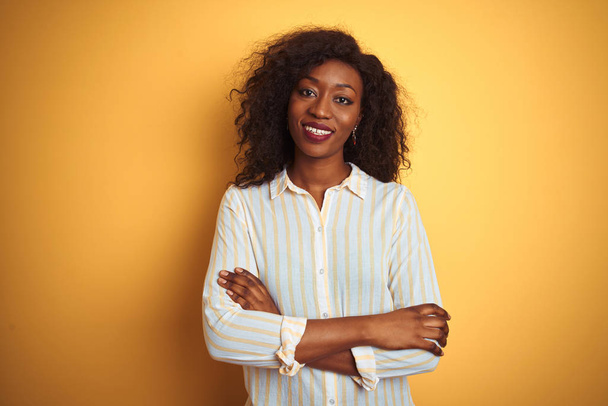 Afro-amerikai nő visel csíkos inget állva az elszigetelt sárga háttér boldog arc mosolyogva keresztkaron nézi a kamerát. Pozitív személy. - Fotó, kép