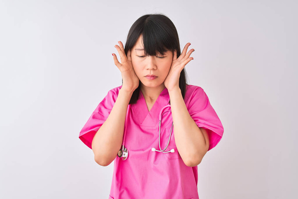 Young beautiful Chinese nurse woman wearing stethoscope over isolated white background with hand on head for pain in head because stress. Suffering migraine. - 写真・画像