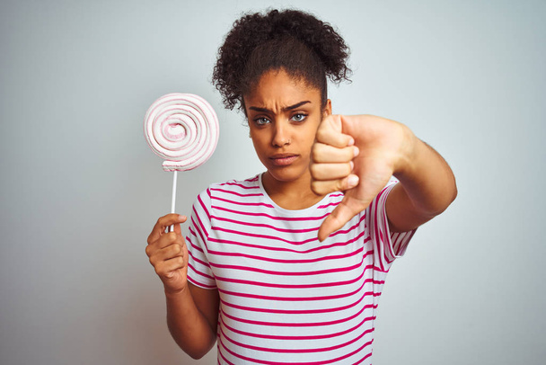 African american teenager woman eating colorful candy over isolated white background with angry face, negative sign showing dislike with thumbs down, rejection concept - Φωτογραφία, εικόνα