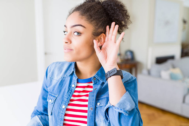 Krásná mladá africká americká žena s afro vlasy nosí ležérní džínová bunda s úsměvem s rukou přes ucho naslouchá slyšení zvěsti nebo drby. Hluchota koncept. - Fotografie, Obrázek