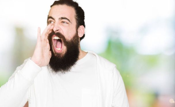 Jonge man met lange haren en baard dragen sportieve sweater schreeuwen en schreeuwen luid naar de andere kant met hand op de mond. Communicatieconcept. - Foto, afbeelding