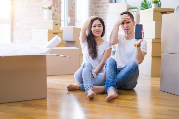 Nádherný pár, který sedí na podlaze, přidržuje smartphone v novém domě kolem lepenkových krabic s rukou na hlavě, šokované hanbou a překvapenou tváří, rozzlobený a frustrovaný. Strach a rozčílený omyl. - Fotografie, Obrázek