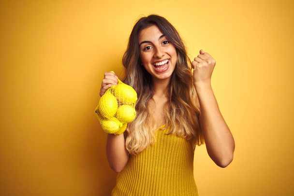 Jeune belle femme tenant filet de citrons sur fond jaune isolé criant fier et célébrant la victoire et le succès très excité, acclamant l'émotion
 - Photo, image
