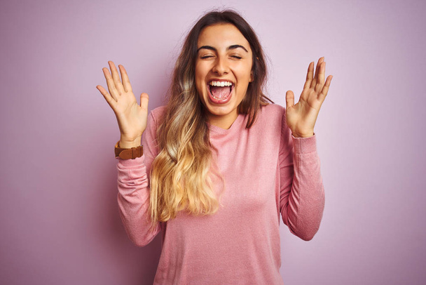 junge schöne Frau in einem Pullover über rosa isoliertem Hintergrund feiert verrückt und verrückt nach Erfolg mit erhobenen Armen und geschlossenen Augen schreit aufgeregt. Siegerkonzept - Foto, Bild