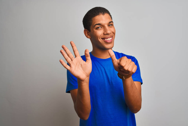 Молодий красивий арабський чоловік у синій футболці стоїть над ізольованим білим тлом, показуючи і вказуючи пальцями номер шість, посміхаючись впевнено і щасливо
. - Фото, зображення