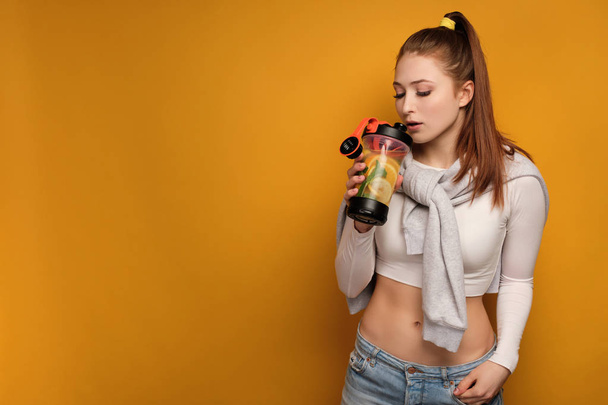 Redhead girl in a short top is standing on a yellow background, going to take a sip from a sports shaker. - Foto, immagini