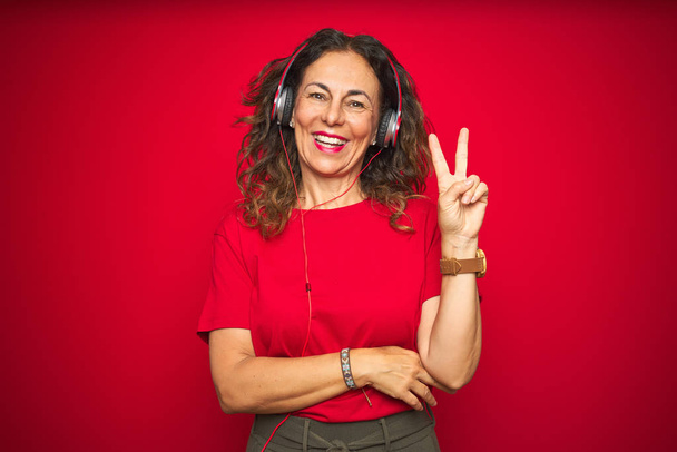 Μεσήλικη γυναίκα που φοράει ακουστικά ακούγοντας μουσική πάνω από κόκκινο απομονωμένο φόντο χαμογελώντας με χαρούμενο πρόσωπο στο μάτι της κάμερας κάνοντας το σημάδι της νίκης. Νούμερο δύο.. - Φωτογραφία, εικόνα