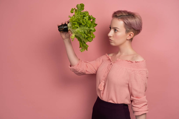 A girl with short pink hair in a blouse looks thoughtfully at a salad in her hand, standing in profile on a pink background. - Foto, Imagem
