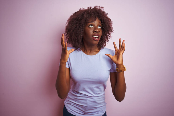 Mladá Africká Afro žena, která nosí tričko stojící nad izolovaným růžovým pozadím bláznivě a bláznivě vykřikuje a křičí s agresivním výrazem a zdviženýma rukama. Koncepce frustrace. - Fotografie, Obrázek