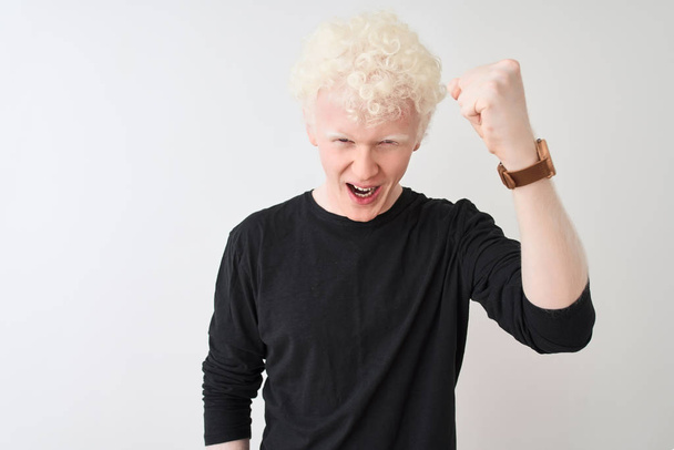 Mladý albín plavovlasý muž na černém tričku, stojící nad izolovaným bílým pozadím, rozzlobený a šílenou pěstí, frustrovaný a rozzuřený, zatímco křičí hněvem. Zuřivost a agresivní koncepce. - Fotografie, Obrázek