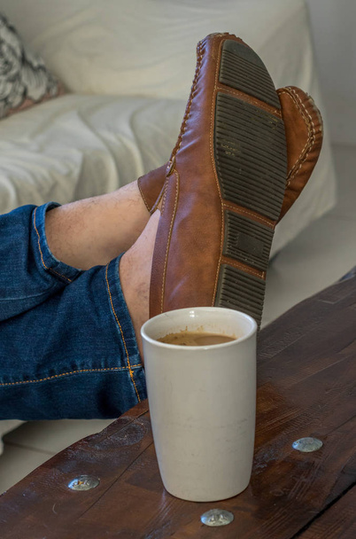 Nohy s koženými botami izolované na stole vedle šálku obrazu kávy ve svislém formátu - Fotografie, Obrázek