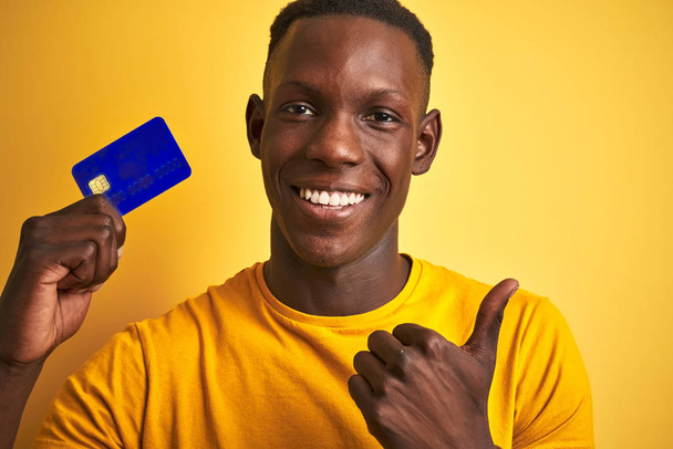 Afrikanisch-amerikanischer Kunde Mann hält Kreditkarte über isoliertem gelben Hintergrund glücklich mit großem Lächeln tun ok Zeichen, Daumen nach oben mit Fingern, ausgezeichnetes Zeichen - Foto, Bild