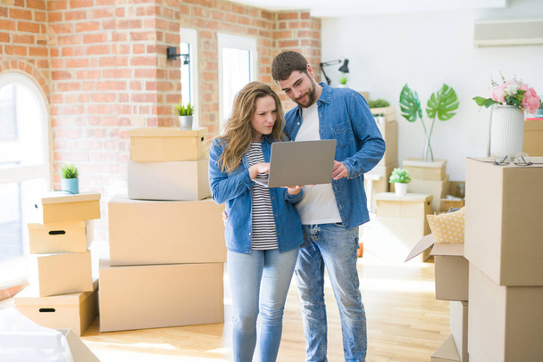 Yeni bir daireye taşımak için mutlu karton kutular etrafında bir odada bilgisayar laptop ayakta kullanarak genç çift - Fotoğraf, Görsel