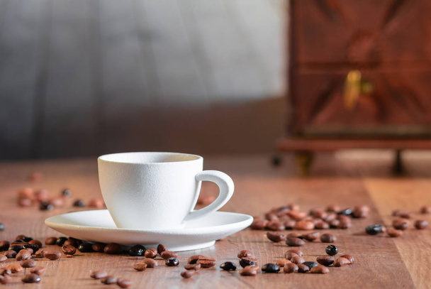 Φωτογραφίες πολλών κόκκων καφέ που περιβάλλουν ένα λευκό πορσελάνινο Κύπελλο με επιλεκτική εστίαση σε ένα ξύλινο τραπέζι. Έννοια της χαλάρωσης και του αρώματος του καφέ. - Φωτογραφία, εικόνα