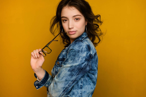 A curly dark-haired girl in a jeans stands on a yellow background in a half-turn and looks at the camera holding glasses in hand - Fotó, kép