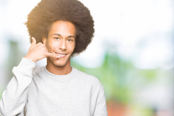 スポーティなスウェットシャツを着たアフロヘアの若いアフリカ系アメリカ人男性は、電話で話すのと同じように手と指で電話ジェスチャーをして微笑んでいます。概念の伝達. - 写真・画像