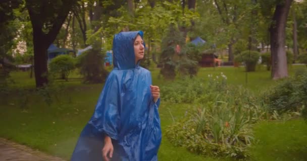 Closeup portrait of young caucasian female in blue raincoat walking in park while it is raining outdoors - Video