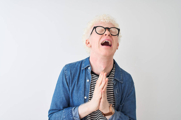 Jonge albino blonde man dragen denim shirt en bril over geïsoleerde witte achtergrond smeken en bidden met de handen samen met hoop uitdrukking op het gezicht zeer emotioneel en bezorgd. Om vergiffenis vragen. Religie concept. - Foto, afbeelding