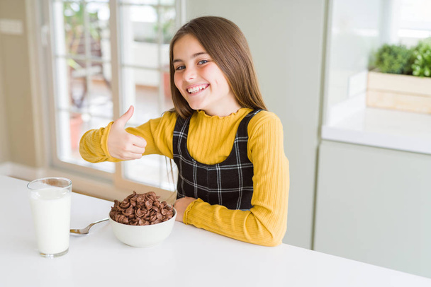 Beautiful young girl kid eating chocolate cereals and glass of milk for breakfast doing happy thumbs up gesture with hand. Approving expression looking at the camera with showing success. - Photo, image