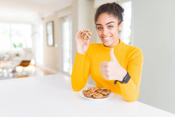 Chica afroamericana joven comiendo galletas de chispas de chocolate como aperitivo dulce feliz con una gran sonrisa haciendo signo de ok, pulgar hacia arriba con los dedos, señal excelente
 - Foto, Imagen