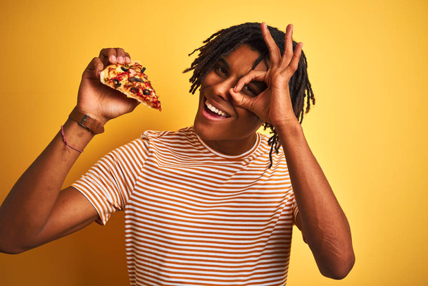 Afro american man with dreadlocks eating slice of pizza over isolated yellow background with happy face smiling doing ok sign with hand on eye looking through fingers - Foto, Bild