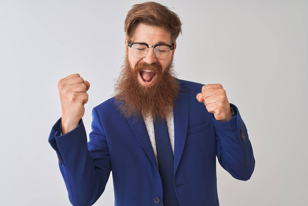 Young redhead irish businessman wearing suit and glasses over isolated white background very happy and excited doing winner gesture with arms raised, smiling and screaming for success. Celebration concept. - Foto, afbeelding
