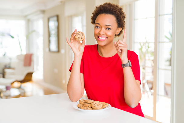 Joven afroamericana americana comiendo galletas fritas de chocolate sorprendida con una idea o pregunta apuntando con el dedo con la cara feliz, número uno
 - Foto, Imagen