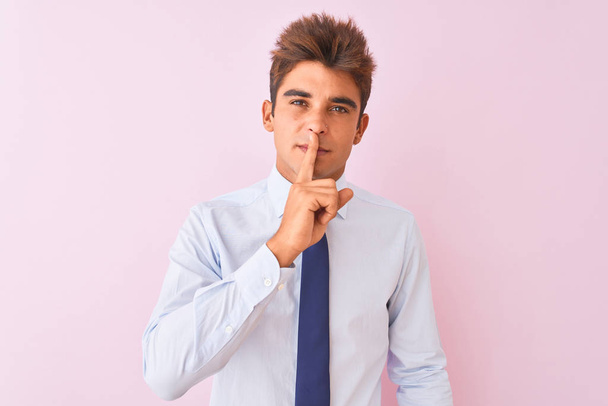 Νέος όμορφος επιχειρηματίας φορώντας πουκάμισο και γραβάτα στέκεται πάνω από απομονωμένο ροζ φόντο ζητώντας να είναι ήσυχος με το δάχτυλο στα χείλη. Σιωπή και μυστική αντίληψη. - Φωτογραφία, εικόνα