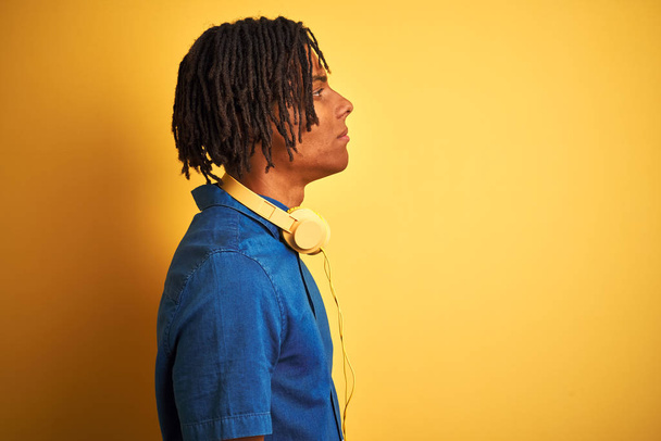 Afro american man with dreadlocks wearing headphones over isolated yellow background looking to side, relax profile pose with natural face with confident smile. - Фото, зображення
