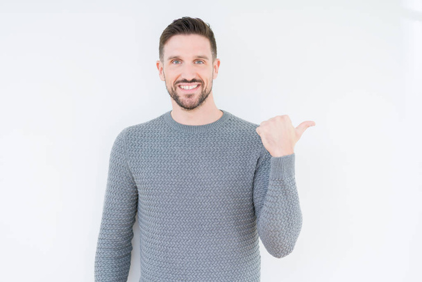 Young handsome man wearing casual sweater over isolated background smiling with happy face looking and pointing to the side with thumb up. - Fotografie, Obrázek