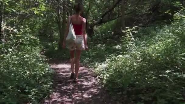 backside view girl in blouse and denim shorts walks along path through green forest. Slowmotions. - Footage, Video